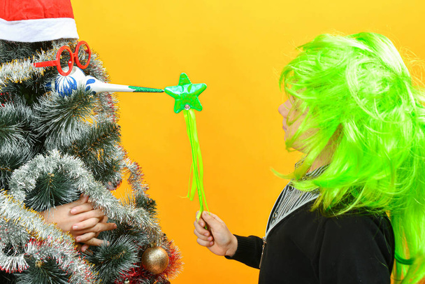 Un garçon avec une perruque verte se tient à côté d'un arbre de Noël avec les mains d'un homme. - Photo, image