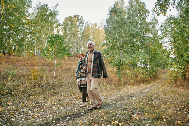 Šťastný starší pár v teplém casualwear pohybující se po silnici nebo lesní cestě - Fotografie, Obrázek