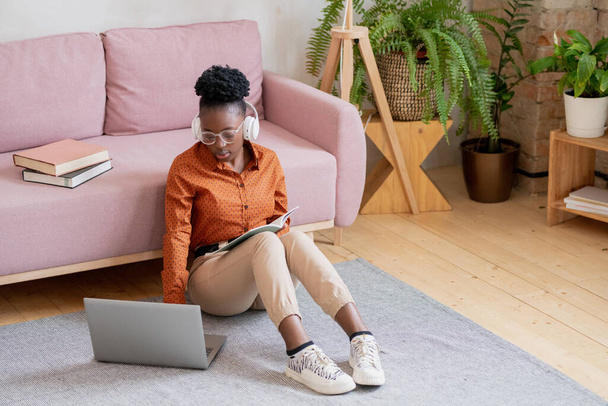 Jeune femme noire sérieuse en casual et écouteurs assis sur le sol devant un ordinateur portable - Photo, image