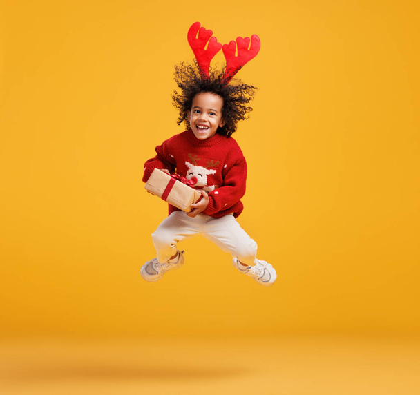 Emocionado niño afroamericano en jersey de Navidad y cuernos de ciervo saltando en el aire con la caja de regalo de Navidad - Foto, imagen