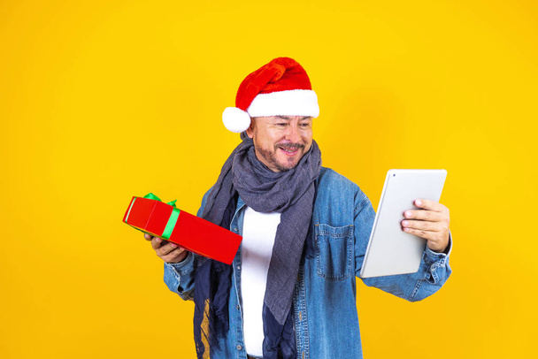 ラテン語のシニア男性がクリスマスビデオ通話を持っている黄色の背景にデジタルタブレットやタッチパッドを使用 - 写真・画像