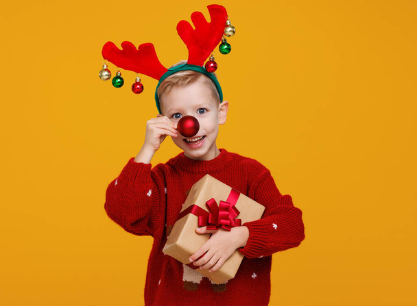 Cheerful little boy with wrapped Christmas gift holding red christmas tree ball in front of his nose - Photo, Image