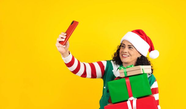 Πορτρέτο της ενήλικης Λατίνας μεσήλικας που κρατά το κινητό τηλέφωνο τραβώντας μια φωτογραφία selfie σε μια χριστουγεννιάτικη ιδέα σε κίτρινο φόντο στο Μεξικό Λατινική Αμερική - Φωτογραφία, εικόνα