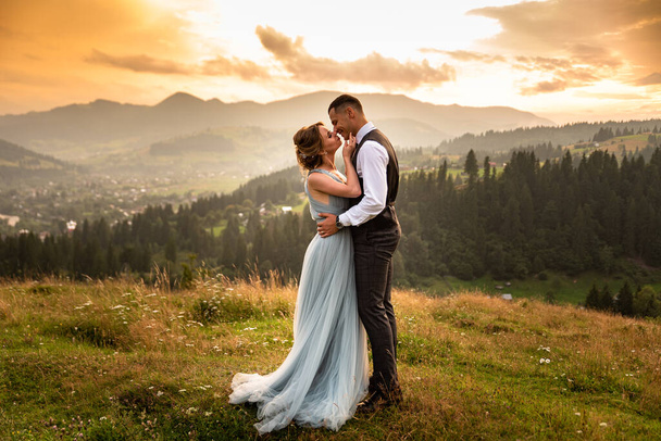 Весілля в горах. Красива пара наречених цілується стоячи на горі на тлі заходу сонця в горах
.  - Фото, зображення