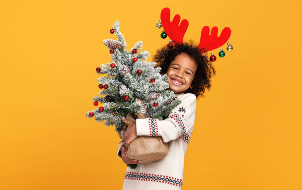Kırmızı geyik boynuzları takmış küçük Afrikalı Amerikalı çocuk elinde karlı Noel ağacı tutuyor. - Fotoğraf, Görsel