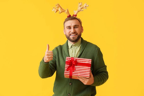 Szczęśliwy młody człowiek z prezentem świątecznym pokazującym gest kciuka do góry na tle koloru - Zdjęcie, obraz