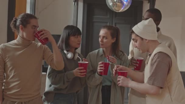 Średnie powolne ujęcie pięciu wesołych wielonarodowych przyjaciół z uniwersytetu podnoszących swoje czerwone kubki przed kamerą podczas zabawy na studenckiej imprezie w kampusie - Materiał filmowy, wideo