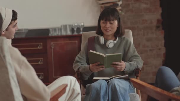 Kézi közepes lassítású felvétel női ázsiai diák olvasás könyv hangosan ül körbe más diákokkal irodalmi klub ülésén campus - Felvétel, videó