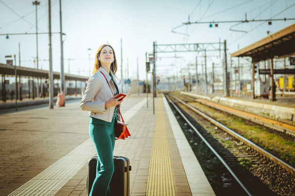 Nő elegáns ruhákat vár a vasútállomáson platform segítségével okostelefon eltölteni az időt. Utazás és üzletasszony fogalma - Fotó, kép