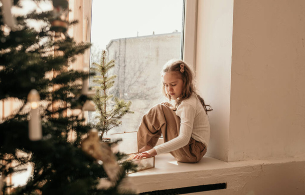 una niña está sentada en el alféizar de la ventana de la habitación y jugando con una caja cerca del árbol de Navidad - Foto, imagen