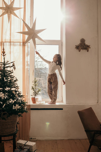 una niña está de pie en el alféizar de la ventana y jugando con una guirnalda estrella - Foto, imagen