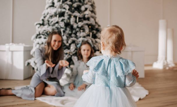 una bambina si alza con la schiena e fa i primi passi verso la madre e la sorella in una stanza con un albero di Natale - Foto, immagini