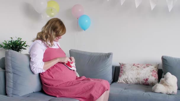 Hymyilevä ja onnellinen raskaana oleva valkoihoinen nainen istuu olohuoneessa katsoessaan pieniä vauvanvaatteita.. - Materiaali, video