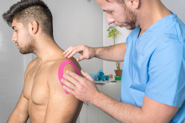Fisioterapeuta aplicando cinta de kinesio en el hombro del paciente masculino. Kinesiología, fisioterapia, concepto de rehabilitación. de cerca - Foto, imagen