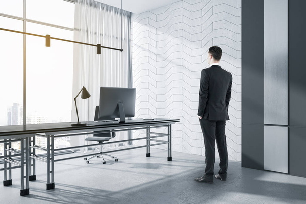 Attractive businessman standing in modern concrete office interior with window and city view, daylight, furniture and various objects. Workplace concept - Photo, image