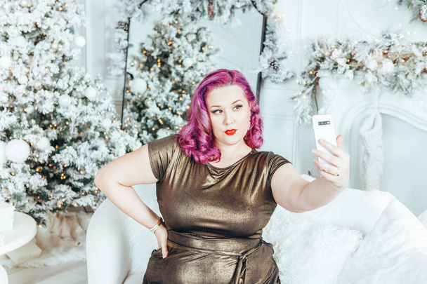 Mooie jonge blanke plus size model vrouw in lange gouden olijf kleur jurk nemen selfie foto portret in vakantie winter decoratie thuis - Foto, afbeelding