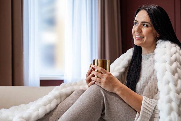 Happy young brunette woman relaxing with mug of hot beverage covered by warm plaid - Φωτογραφία, εικόνα