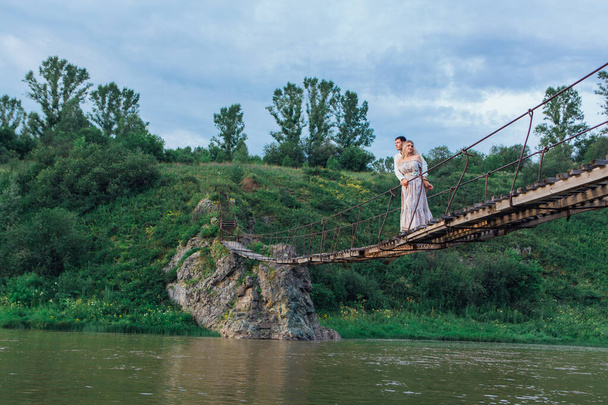 Beautiful young romantic couple standing and hugging on the suspension bridge above the river - Фото, зображення
