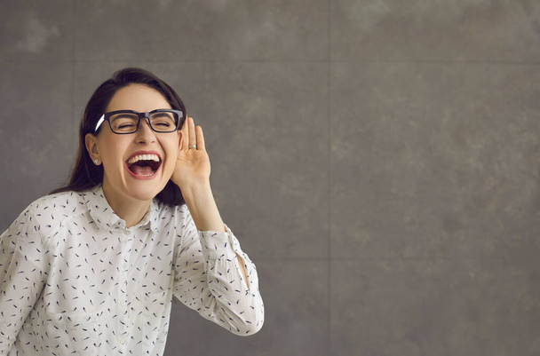 Happy woman with hand at ear listening to funny news on gray copy space background - Фото, изображение