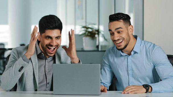 Zwei arabische junge männliche Geschäftsleute, die mit Laptop im modernen Büro sitzen und mit dem Verhandlungsergebnis zufrieden sind, nachdem sie einen Vertrag abgeschlossen haben, die indischen Kollegen erhalten gute Nachrichten und fühlen sich dankbar - Foto, Bild