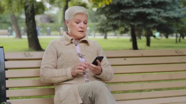 Atractiva mujer mayor de pelo gris de la edad de jubilación sentada en el banco del parque haciendo videollamada, mirando la cámara del teléfono inteligente. Abuela jubilado utiliza moderno gadget internet hablando en el teléfono móvil al aire libre - Imágenes, Vídeo