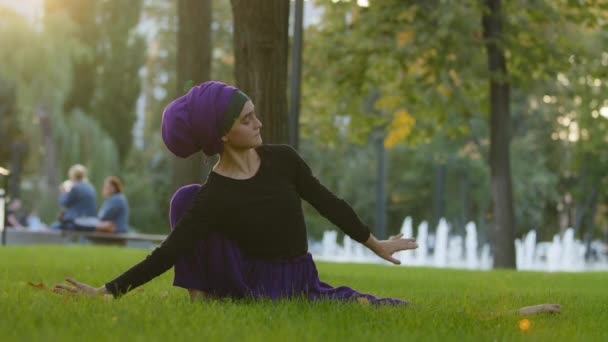 Muslimi tyttö hijabissa islamilainen nainen naispuolinen joogi urheilija harjoitellaan jooga harjoitus puistossa vihreällä nurmikolla istuu narussa yhdellä jalalla mutka ulottuu ylös tekemässä pitoa takana venyttely joustavuutta - Materiaali, video