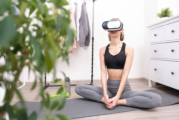 A girl in virtual reality glasses is engaged in yoga and fitness remotely. Future technology concept. A young woman goes in for sports at home online. - Fotó, kép