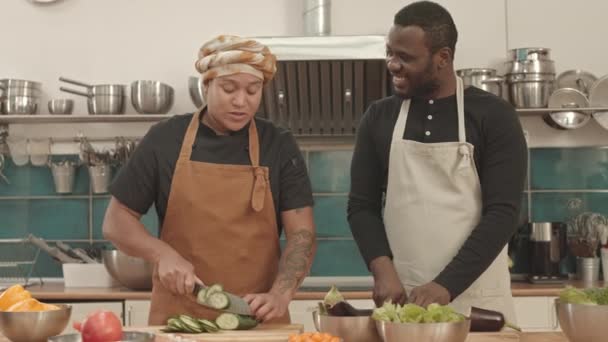 Средняя юная афроамериканка и мужчина в фартуках, стоят на острове на кухне, режут свежий огурец и баклажаны на досках, разговаривают и улыбаются - Кадры, видео