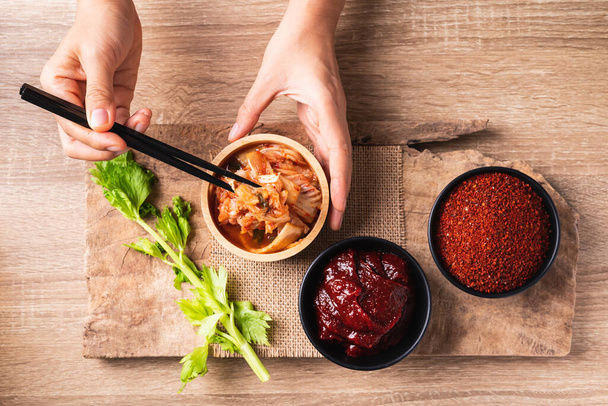 Koreański kimchi kapusta jedzenia za pomocą pałeczek z gochujang (czerwona pasta chili) i chili w proszku w misce na drewnianym tle, Widok na blat stołu - Zdjęcie, obraz