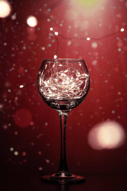 Transparent wine glass, with shining lights of bright bulbs and a beautiful background. A shining glass with bright lights creates an atmosphere of holidays and a desire to celebrate Christmas and New Year. - Foto, Imagem