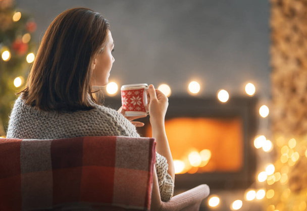 Young woman is drinking coffee near Christmas tree at home. - Φωτογραφία, εικόνα