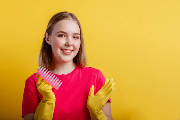 Γυναίκα Πορτρέτο με βούρτσα καθαρισμού σε λαστιχένια γάντια. Νεαρή ξανθιά χαρούμενη χαμογελαστή γυναίκα έτοιμη να καθαρίσει το σπίτι με οικιακές προμήθειες απομονωμένες σε κίτρινο φόντο. Αντιγραφή χώρου - Φωτογραφία, εικόνα