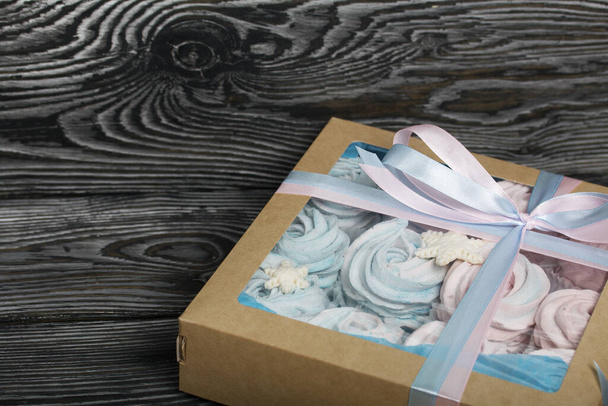 Marshmallow blue and pink. Lies in craft packaging. Decorated with mastic snowflakes. The box is tied with a ribbon tied to a bow. On black pine boards. - Photo, image