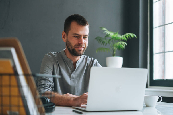 Jeune homme d'affaires souriant à manches longues gris caduc travaillant à un ordinateur portable dans une pièce de bureau moderne sombre - Photo, image