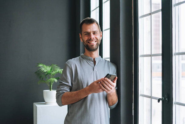Jeune homme barbu à manches longues grises avec smartphone dans les mains lisant le message debout à la fenêtre dans le bureau moderne avec des murs sombres - Photo, image