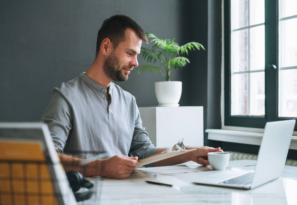 Молодий усміхнений бізнесмен у випадковому сірому рукаві працює в ноутбуці в темній сучасній офісній кімнаті
 - Фото, зображення