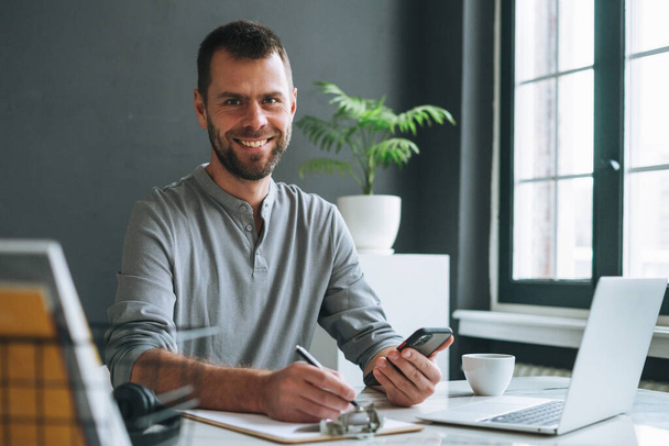 Joven hombre de negocios sonriente en manga larga gris informal que trabaja en el ordenador portátil en la sala de oficina moderna oscura - Foto, Imagen