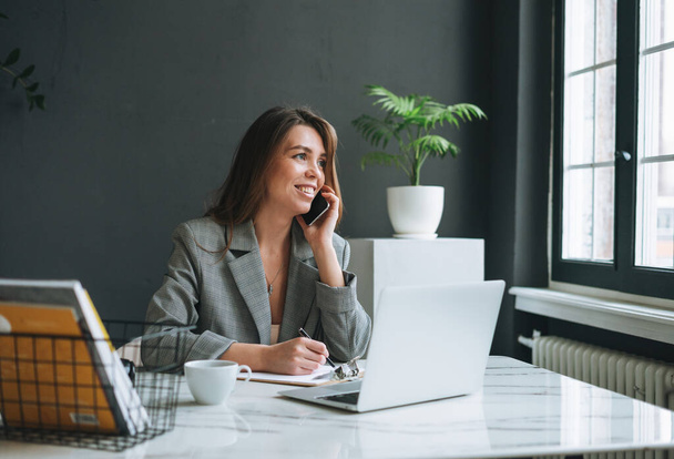 Jovem mulher morena sorridente com cabelos longos em terno cinza usando telefone celular trabalhando no laptop no escritório moderno - Foto, Imagem