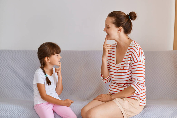 Portrait of woman and little girl having lesson for improving sounds or language difficulties, female child sitting indoor on comfortable sofa and practicing articulation. - Photo, Image