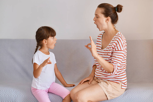 Indoor shot of female physiotherapist posing with small child girl indoors while sitting on sofa, fix speech problems, demonstrates how to pronounce sounds. - Valokuva, kuva