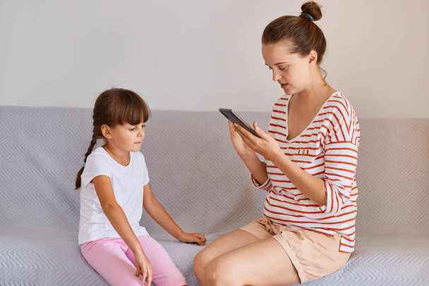 Professional female physiotherapist working with small child girl, sitting on sofa with mobile phone in hands, finds new online lesson in her gadget for training. - Fotografie, Obrázek