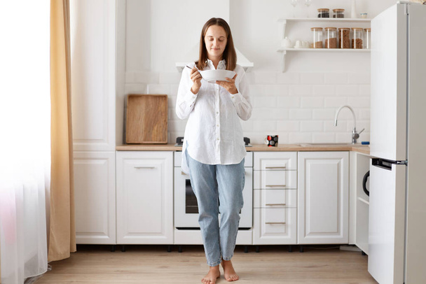 Portrait of happy young housewife eating soup, holding plate in hands while standing against while kitchen set, wearing jeans and shier, having breakfast before going to work. - Foto, imagen