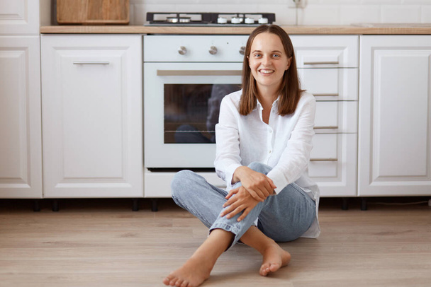 Indoor shot of smiling female with dark hair sitting on floor in kitchen and looking at camera with positive expression, wearing white shirt and jeans. - Fotografie, Obrázek