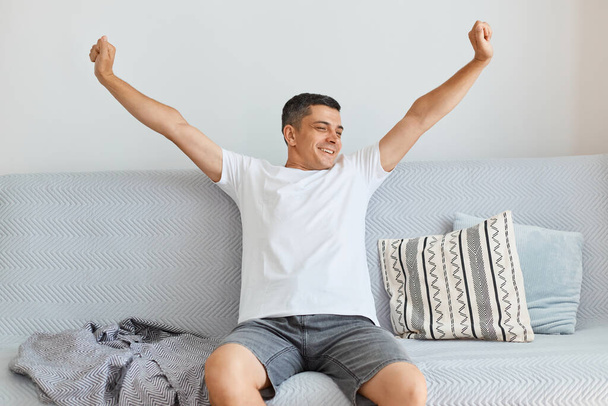 Indoor shot of happy positive attractive brunette man wearing white shirt and jeans, sitting on gray couch in light living room, stretching arms, just wake up in high spirit. - Foto, Imagem