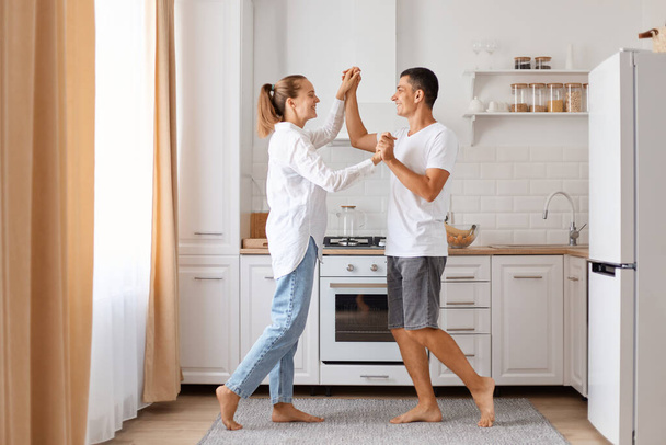 Indoor shot of happy beautiful couple smiling while dancing in kitchen at home, holding each other hands, enjoying spending time together, family wearing white shirts and jeans. - Фото, изображение