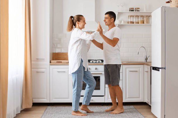 Indoor full length shot of positive couple, young adult man and woman wearing white shirts and jeans, dancing together at home against light kitchen set. - Fotografie, Obrázek