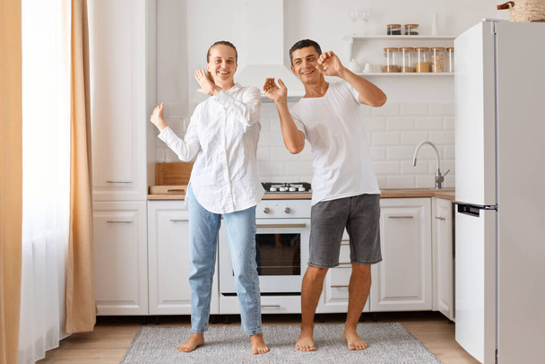 Indoor shot of happy positive optimistic couple dancing happily indoor against white kitchen set, family wearing casual style clothing, raised arms, having fun at weekend. - Foto, Imagem