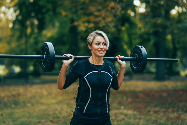 Uma mulher madura em forma está fazendo exercícios de agachamento, vestida com um terno preto com um simulador eletrônico EMS para estimular seus músculos. - Foto, Imagem