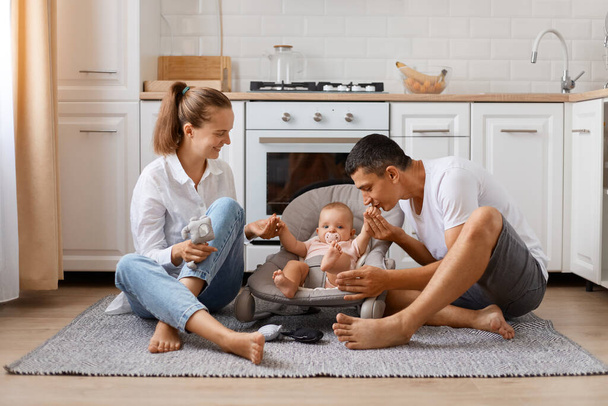 Beautiful Caucasian family father, mother and daughter sitting on floor at cozy white kitchen, loving parents enjoying time with their cute little child, father kissing child's hand. - Foto, Imagem
