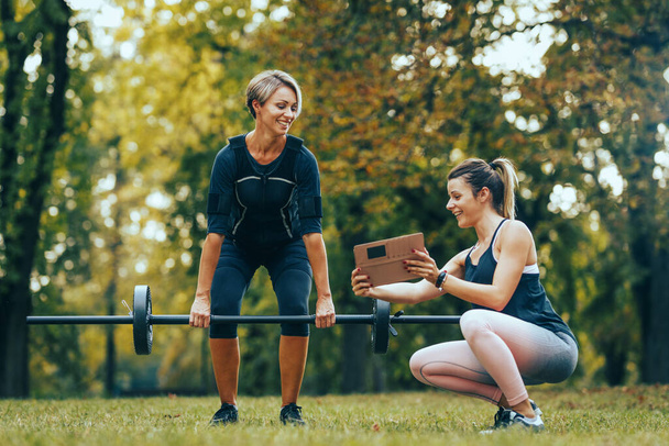 A fit mature woman is doing exercises with personal trainer in the park, dressed in a black suit with an EMS electronic simulator to stimulate her muscles. - Photo, Image